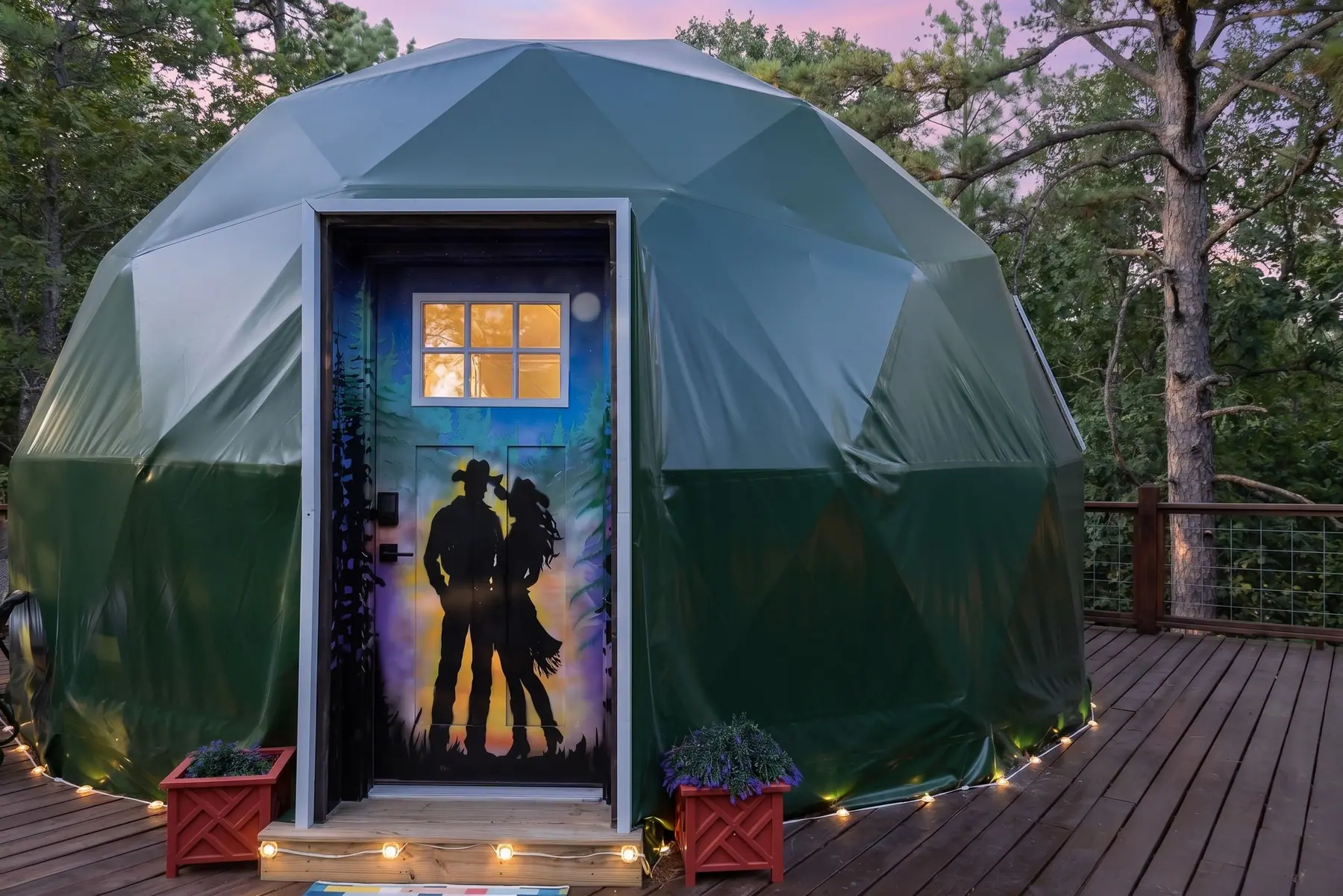 The geodesic glamping dome at dusk, its painted space-cowboy door lit and string lights along the deck